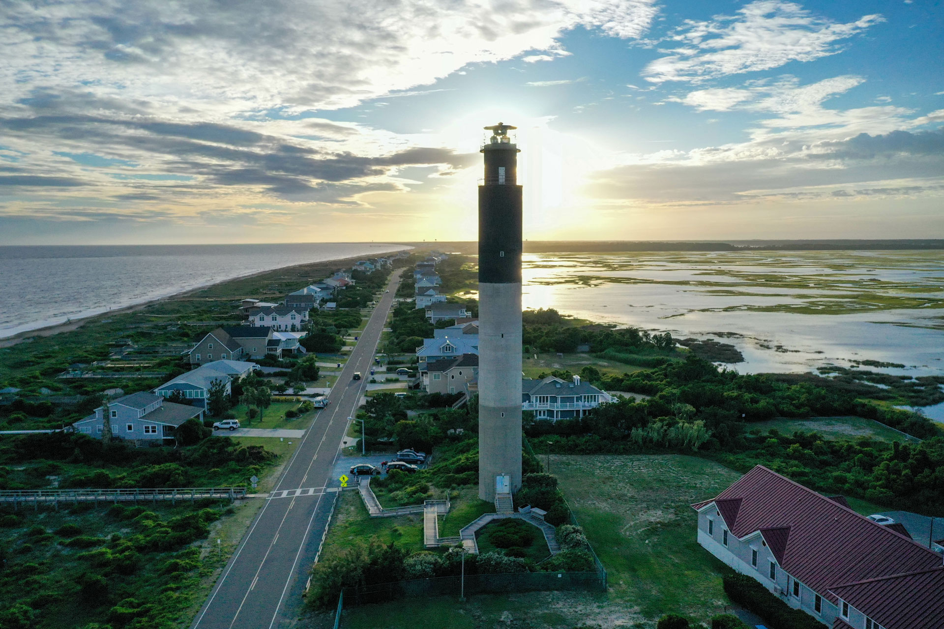 The sun is setting behind the Oak Island, NC lighthouse, surrounded by the ocean and waterway on a partly cloudy day